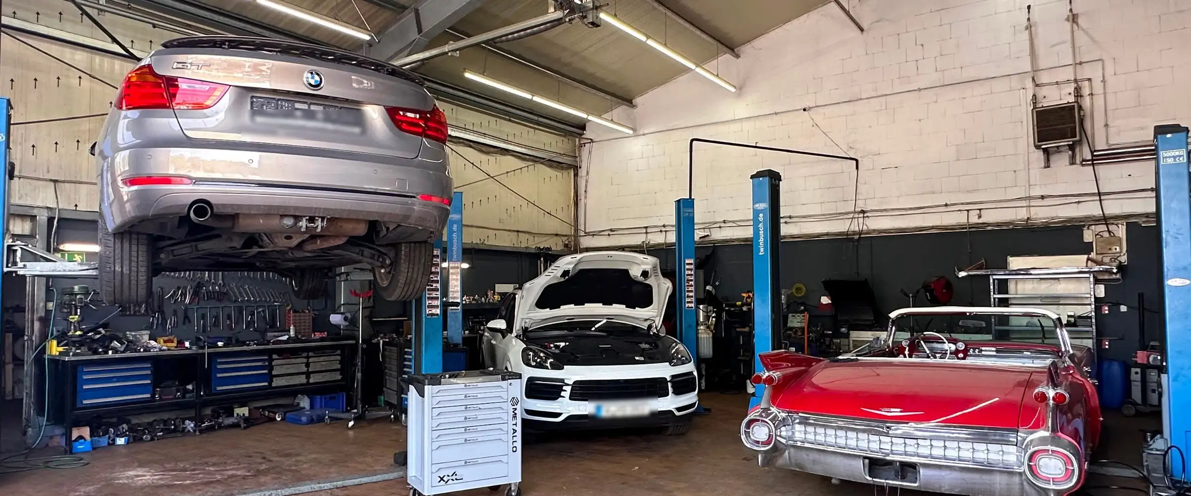 Blick in die Werkstatthalle von Boostwerk Koeln: links ein Kombi angehoben auf der Zwei-Saeulen-Hebebuehne, dahinter die Werkbank, in der Mitte ein SUV mit offener Motorhaube, rechts ein rotes Oldtimer-Cabriolet.