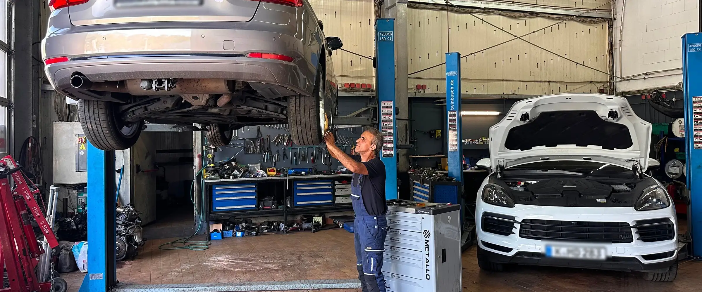 Ein Mechaniker steht unter dem angehobenen Kombi auf der Zwei-Saeulen-Hebebuehne und arbeitet mit einer Handlampe am Unterboden; rechts wartet ein SUV mit offener Motorhaube auf der zweiten Buehne.