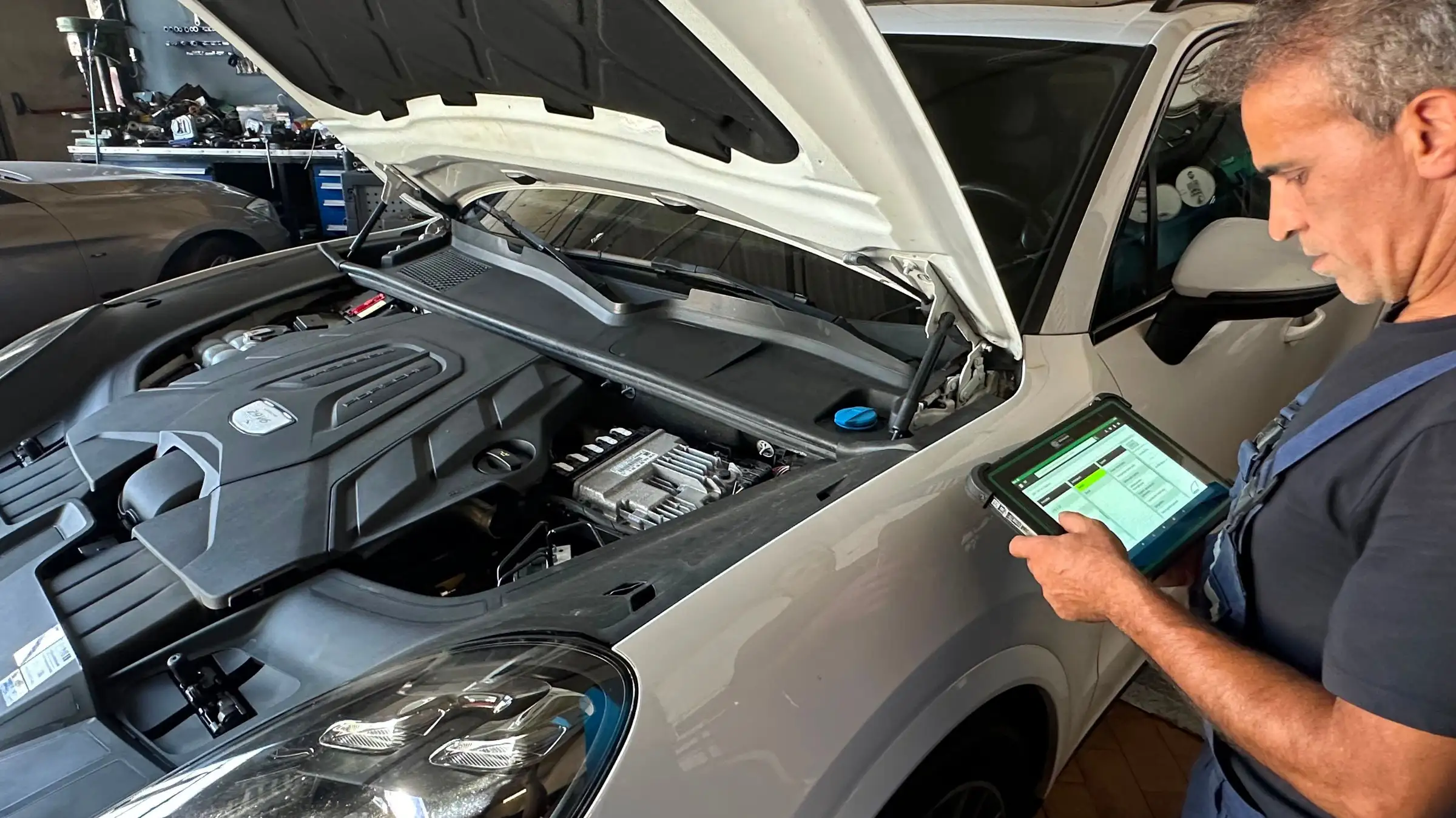 Ein Mechaniker steht am geoeffneten Motorraum eines SUV und liest mit einem Diagnosegeraet den Fehlerspeicher aus; im Motorraum sind Motorabdeckung, Steuergeraet und Kabelbaum zu sehen.