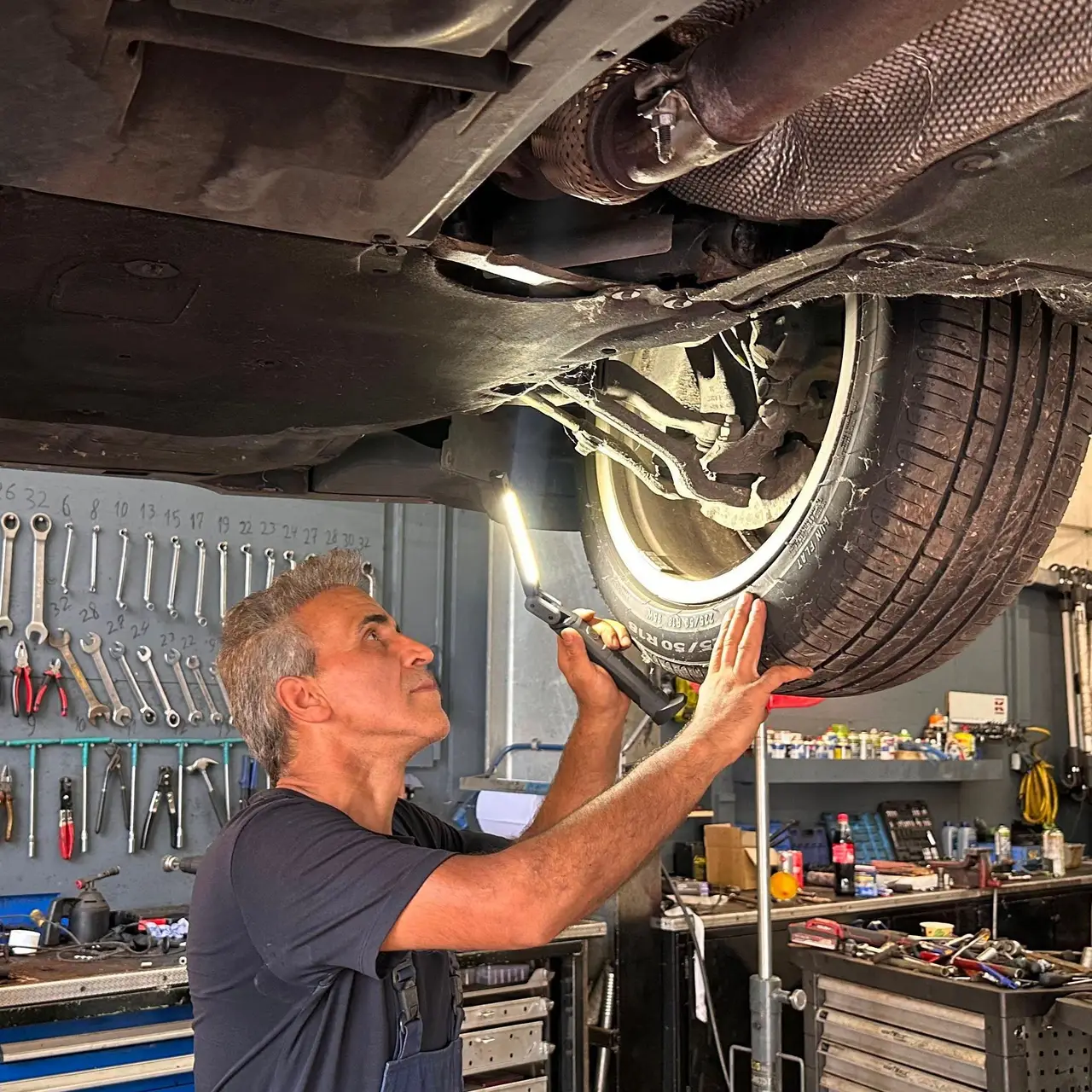 Ein Mechaniker leuchtet mit einer Handlampe unter das angehobene Fahrzeug und prueft Rad und Radaufhaengung; im Hintergrund die Werkzeugwand.